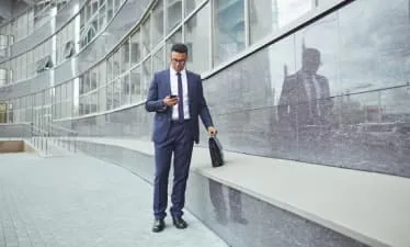 Homme en costume consultant son téléphone devant une façade vitrée, avec une sacoche.